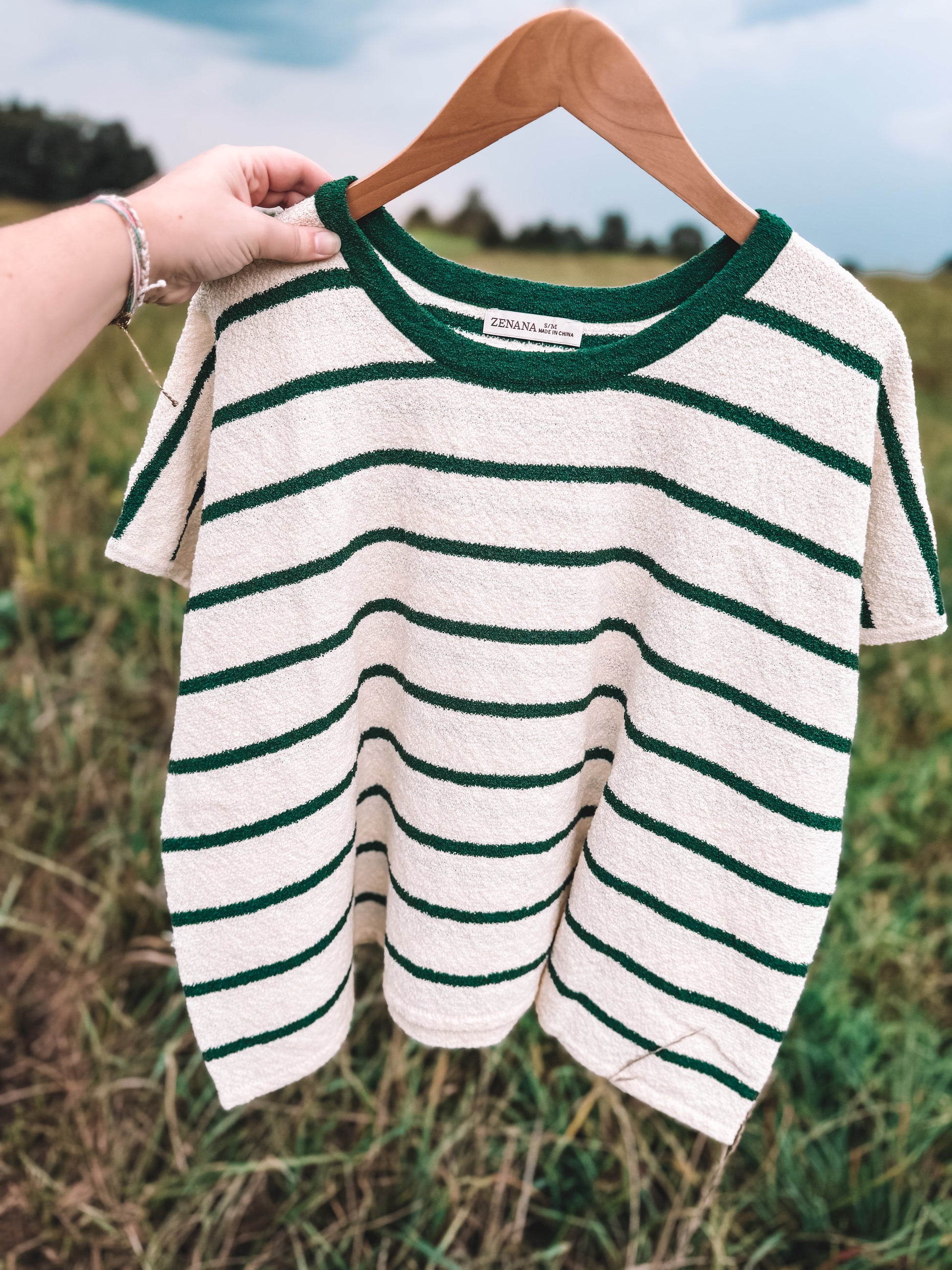 A short-sleeve knit top with dark green horizontal stripes on an off-white base, hanging on a wooden hanger held by a person's hand against an outdoor grassy background.