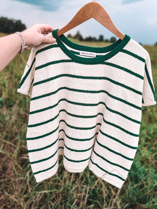 A short-sleeve knit top with dark green horizontal stripes on an off-white base, hanging on a wooden hanger held by a person's hand against an outdoor grassy background.
