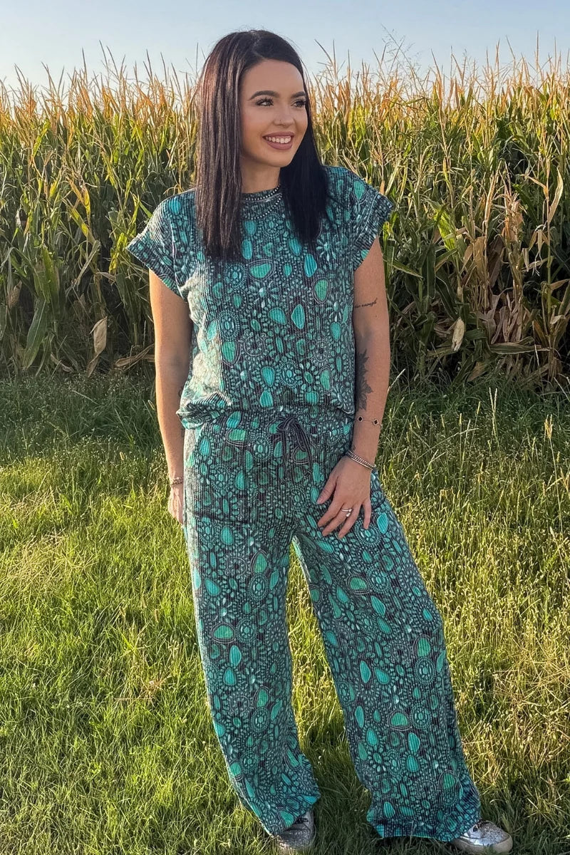 A person standing outdoors in a grassy field wearing loose, wide-leg pants with an allover teal, green, and dark gray paisley-style print, along with a matching top and sneakers.