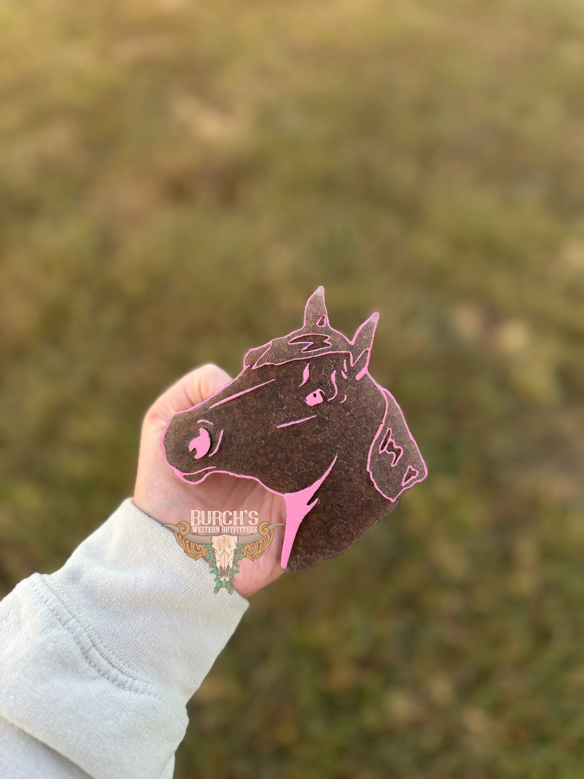 A hand wearing a light gray sweatshirt holds a dark brown felt horse head ornament outlined in pink, with a blurred grassy background.