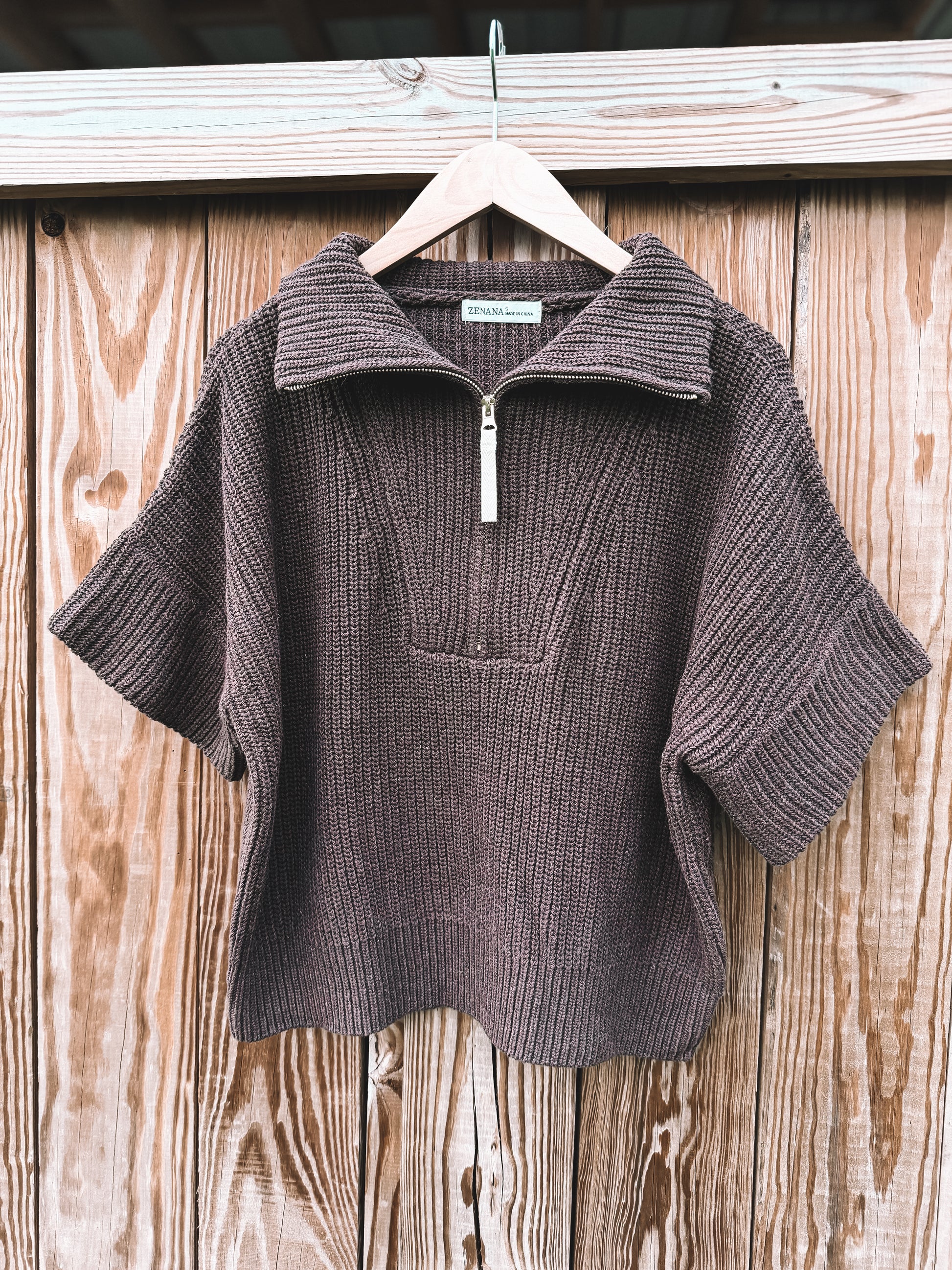 A brown ribbed knit short-sleeve quarter-zip sweater with a mock neck, hanging on a wooden hanger against a rustic wooden plank wall.
