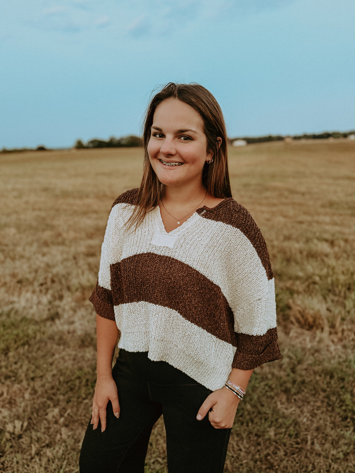 A woman wearing a brown and cream striped knit poncho sweater, standing in a dry grass field under a blue sky.