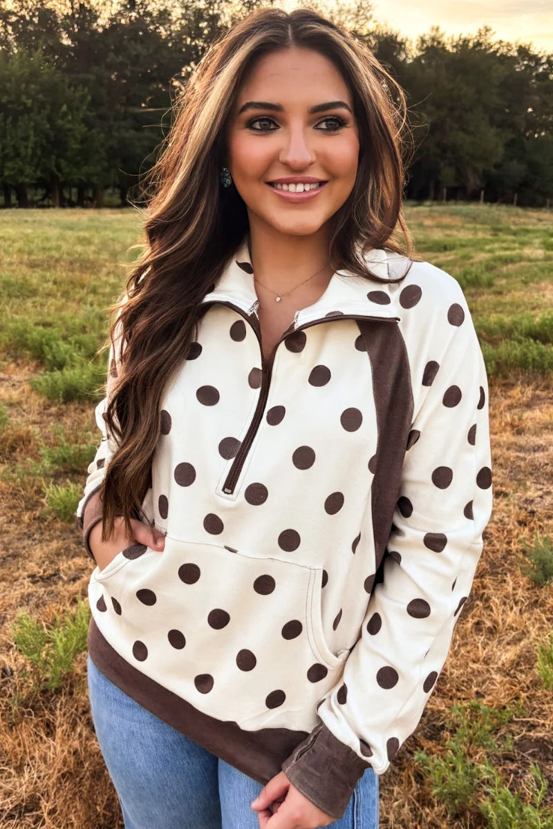 A woman viewed from the front wearing a cream-colored long-sleeve top with dark brown polka dots and a solid dark brown lower panel, standing in an outdoor field at sunset.