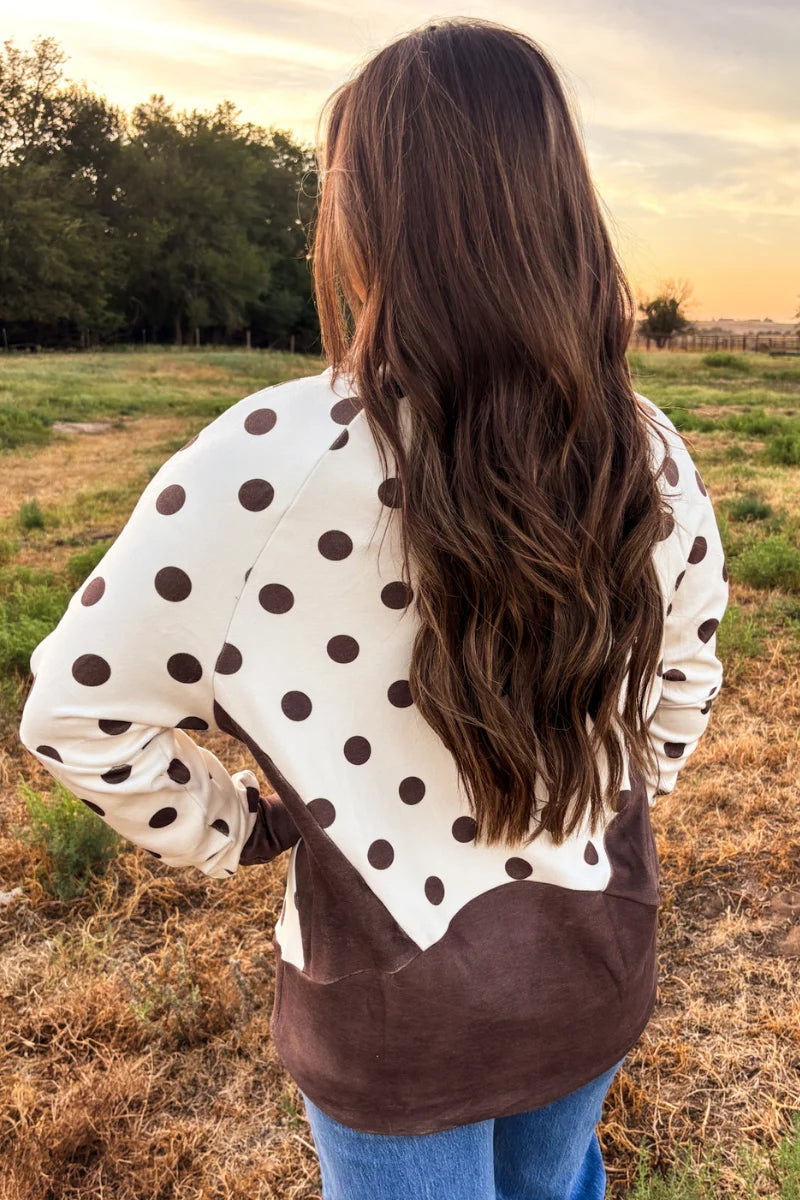 A woman viewed from behind wearing a cream-colored long-sleeve top with dark brown polka dots and a solid dark brown lower panel, standing in an outdoor field at sunset.