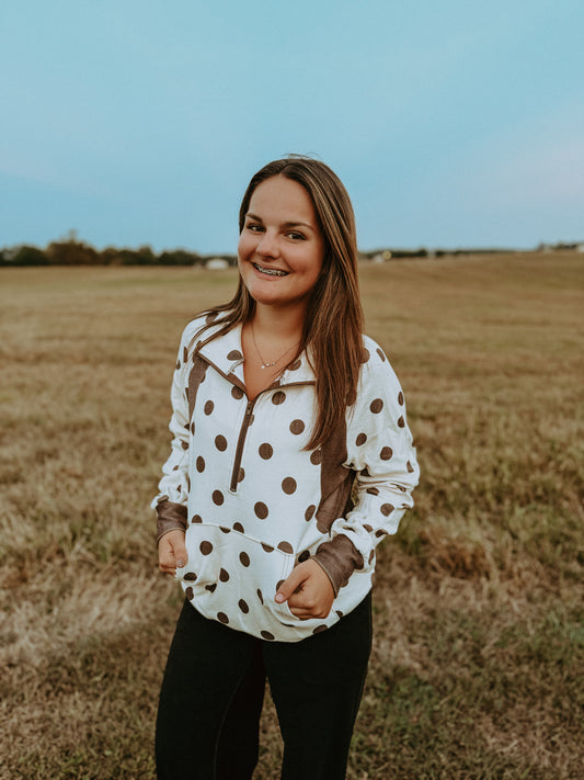 A woman smiling in an outdoor field, wearing a white long-sleeve quarter-zip pullover with brown polka dots and brown trim, paired with black pants.