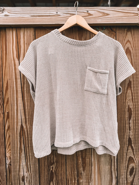 A light beige waffle-knit short-sleeve t-shirt with a chest pocket, hanging on a wooden hanger against a rustic wooden fence background.
