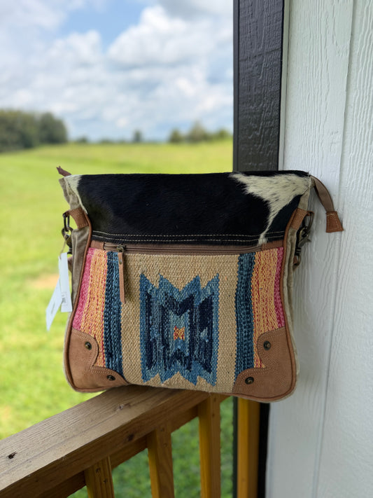 A western-style shoulder bag with a black and white cowhide top flap, tan leather trim, and a colorful woven geometric pattern in blue, pink, and beige, hanging on a wooden porch railing with a green field and cloudy sky in the background.