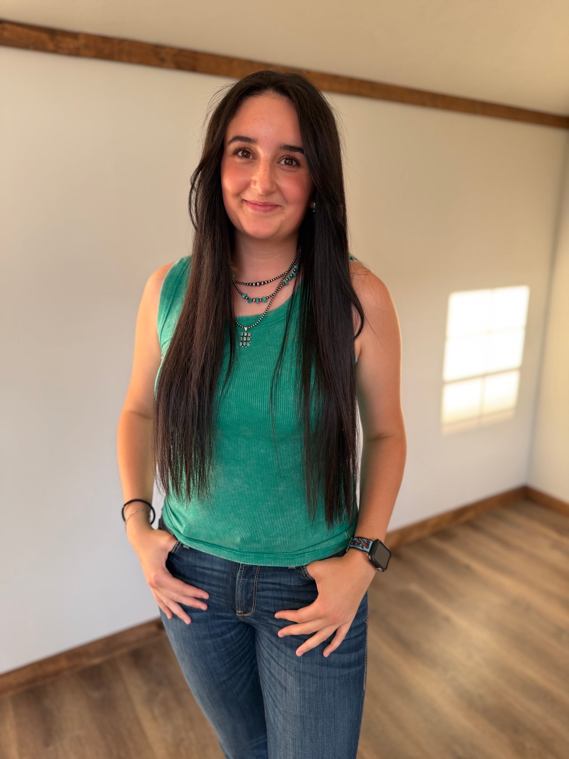 Woman wearing a green tank top and blue jeans standing in a room with wooden flooring and white walls.