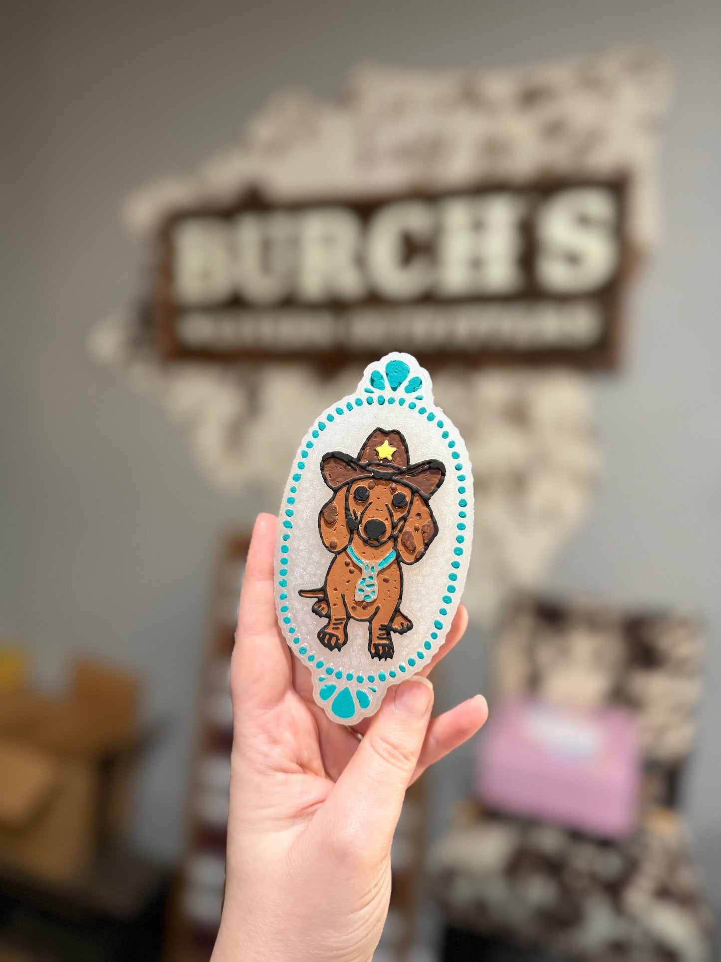Hand holding a sticker of a dachshund in a cowboy hat with 'Burch's' in the background.