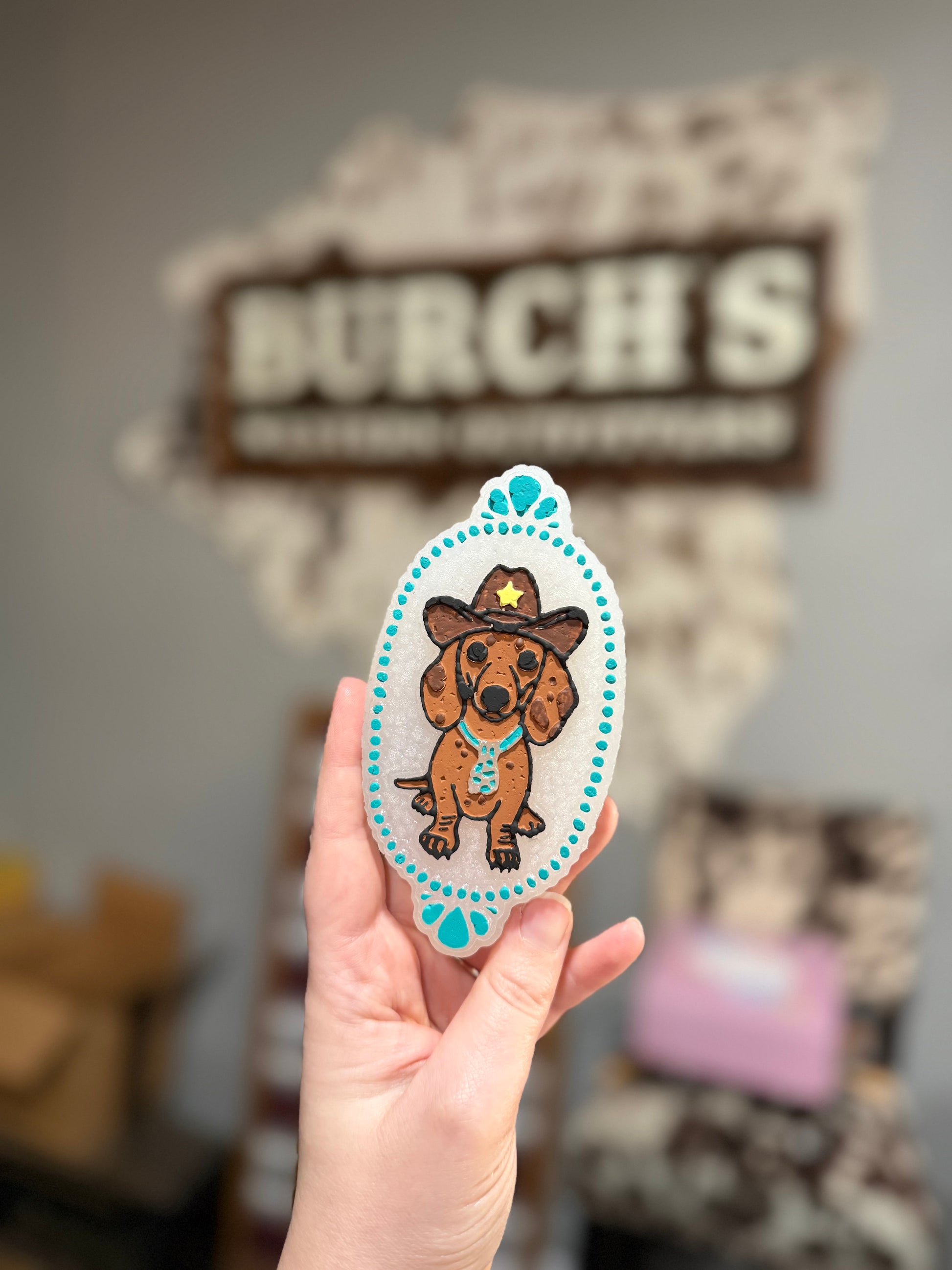 Hand holding a sticker of a dachshund in a cowboy hat with 'Burch's' in the background.