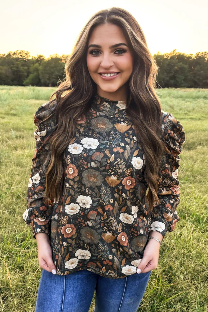 Woman modeling a black floral long-sleeve blouse with puff sleeves and a warm-toned vintage floral print, paired with blue jeans outdoors.
