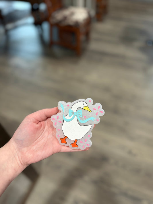 Hand holding a sticker of a cartoon duck with a bow tie on a blurred indoor background