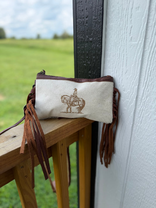 A small beige fabric pouch with dark brown leather fringe and a brown cowboy-on-horse graphic, resting on a wooden porch railing next to a white wall.