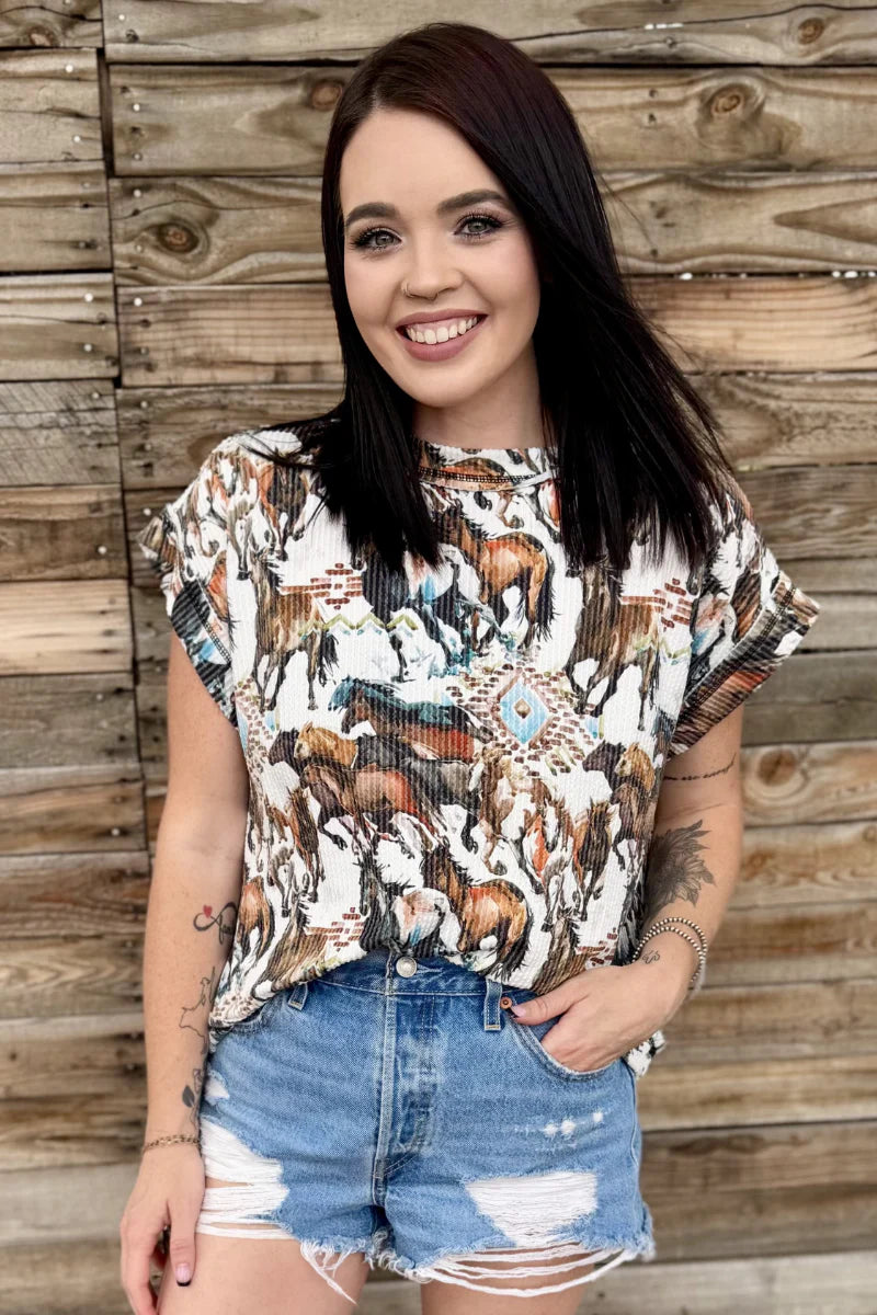 Woman wearing a patterned top and denim shorts standing against a wooden wall.