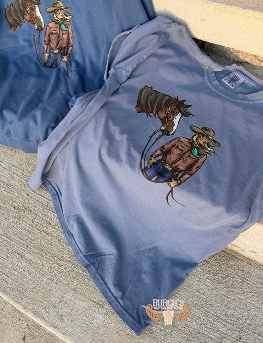 A dusty blue graphic t-shirt laid flat on concrete steps, featuring a western cowboy and horse illustration on the front. A second matching blue t-shirt is partially visible in the background.