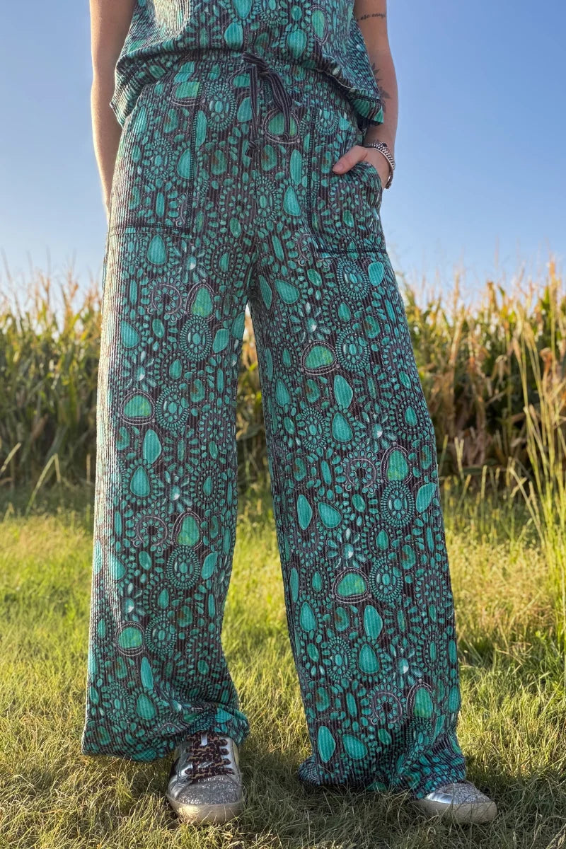 A person standing outdoors in a grassy field wearing loose, wide-leg pants with an allover teal, green, and dark gray paisley-style print, along with a matching top and sneakers.