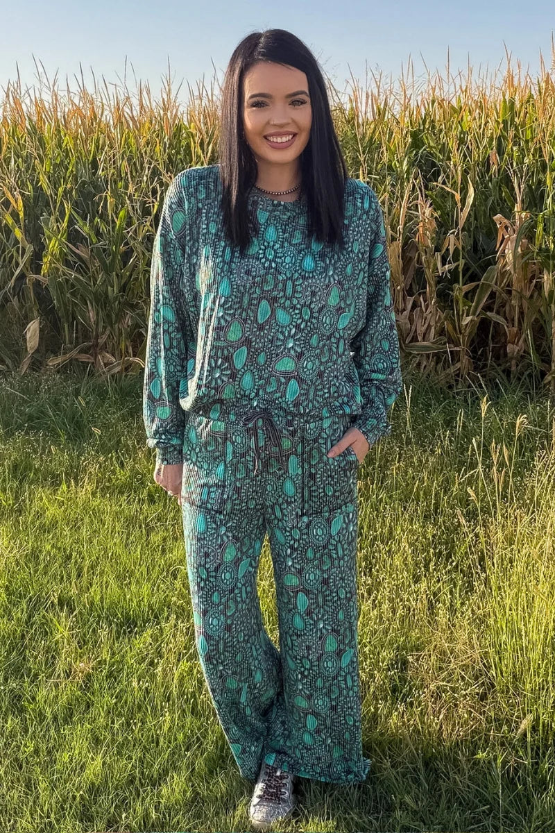 Woman modeling a matching turquoise and charcoal Western print lounge set with a long-sleeve top and wide-leg drawstring pants in front of a cornfield.