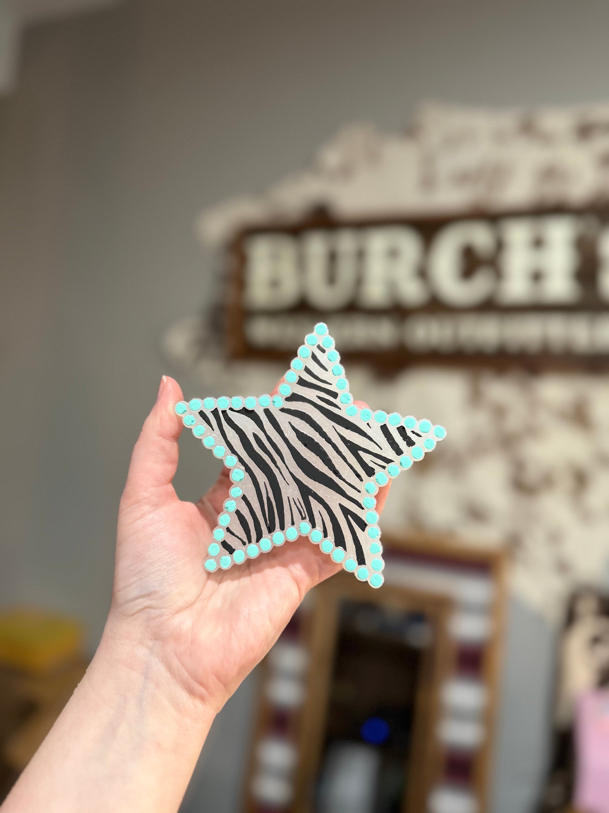 Hand holding a star-shaped object with zebra pattern and turquoise border in front of a blurred background with 'Burch' branding.