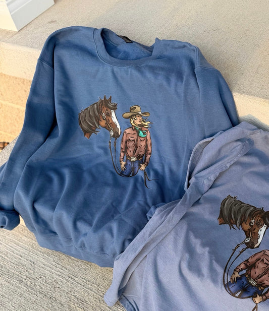 A dusty blue crewneck sweatshirt featuring a graphic print of a cowboy holding a lasso next to a horse, styled on a light beige concrete surface with another folded sweatshirt partially visible.