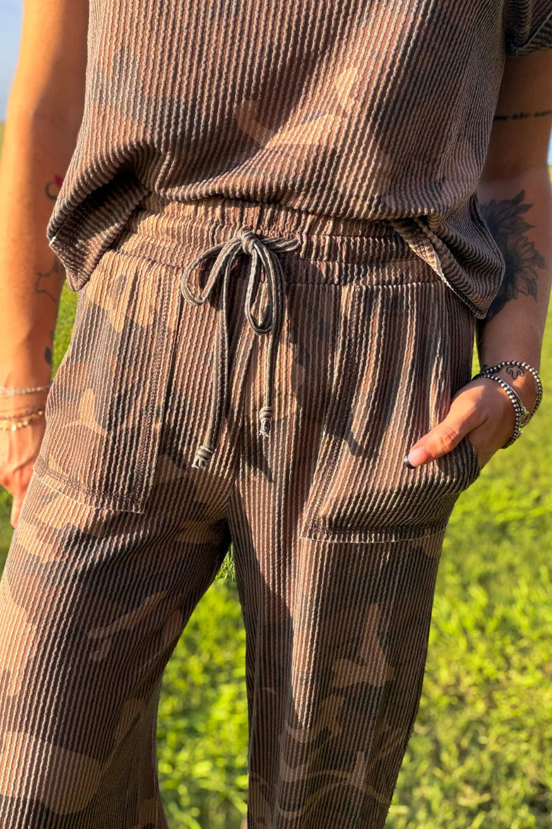 A close-up of a person wearing brown corduroy pants with a subtle camouflage pattern, featuring an elastic drawstring waist and side pockets, set against a blurred green grass background.
