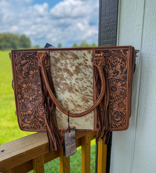 A western-style tote bag with tooled brown leather sides, a cowhide center panel, and long leather fringe, photographed leaning against a wooden railing outdoors with a grassy field and cloudy sky in the background.