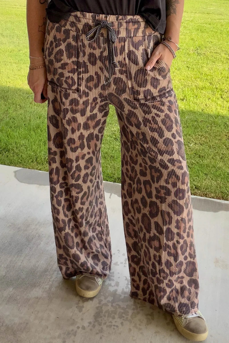 A person wearing wide-leg leopard print pants with a drawstring waist, standing outdoors on a concrete surface with grass in the background.