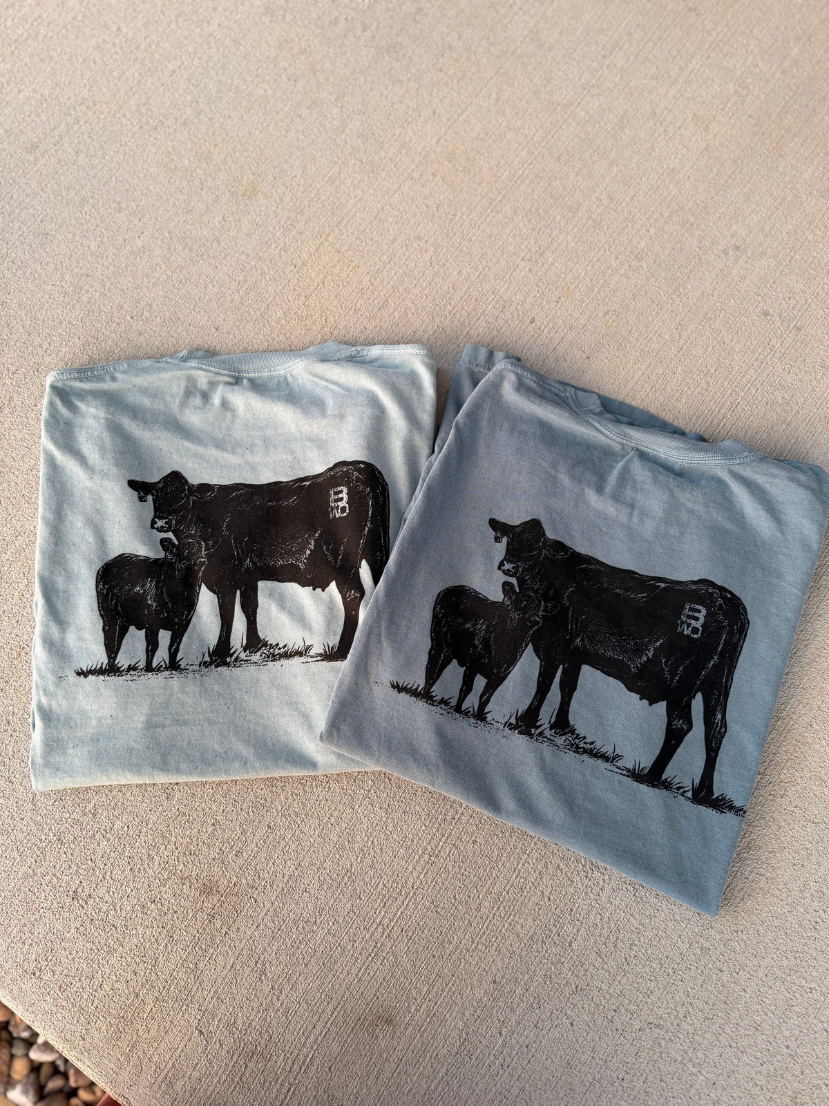 Two folded light blue t-shirts with a black cow and calf graphic print, displayed on a beige textured surface.