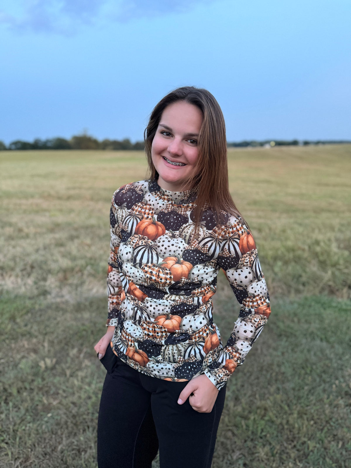 A woman wearing a long-sleeve mock neck shirt with an all-over autumn pumpkin and gourd pattern, standing in an open grassy field under a blue sky.