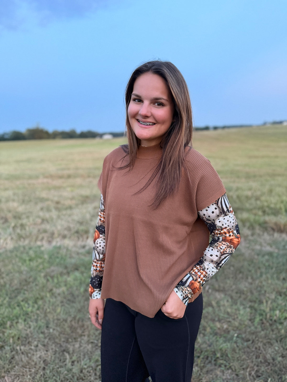 A woman standing in an open grassy field wearing a brown ribbed short-sleeve top with patterned long sleeves and black pants.