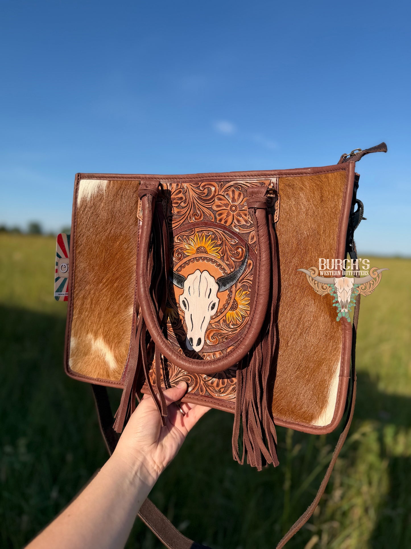 Brown leather handbag with cow design held in front of a grassy field
