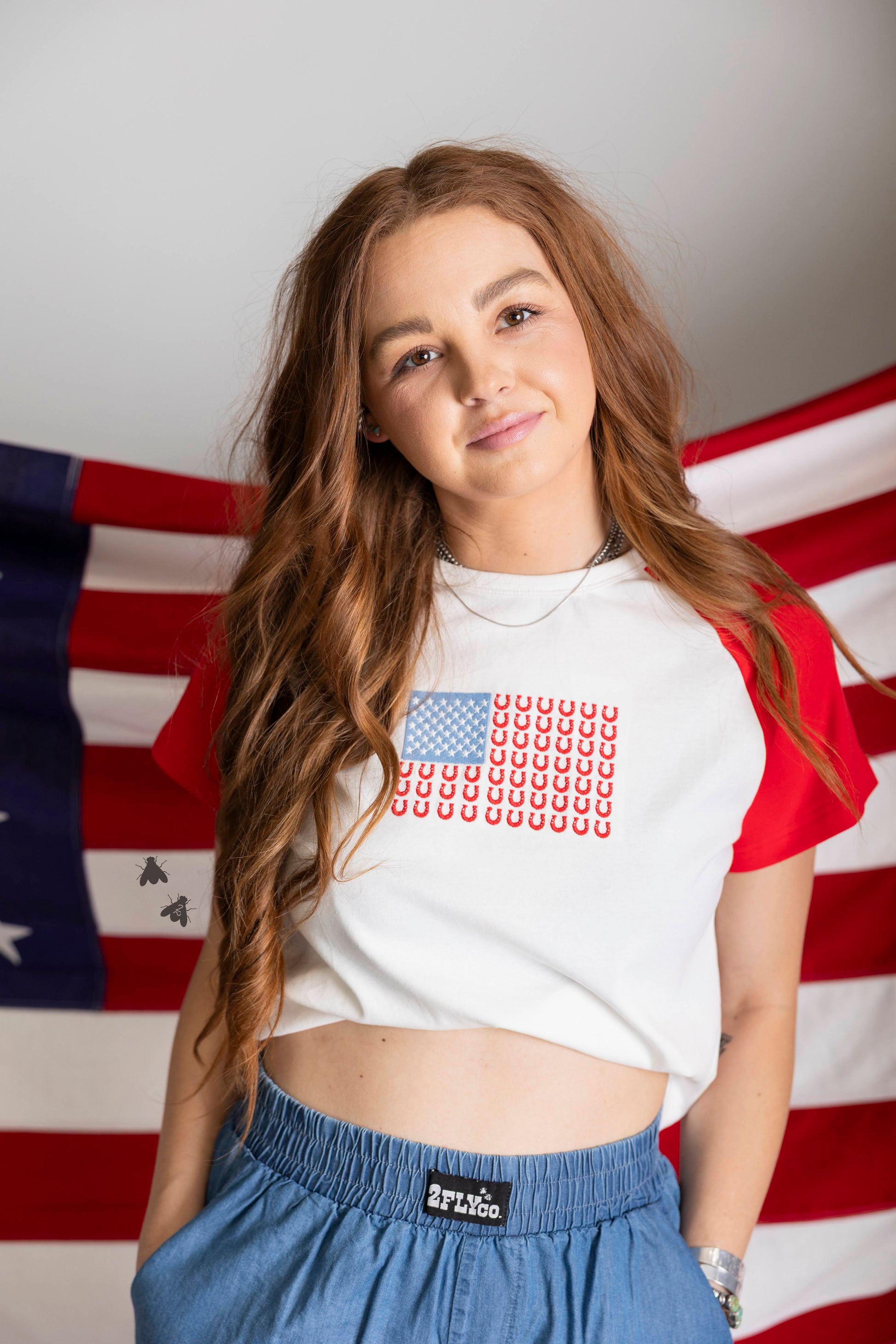 Person wearing a white crop top with a red, white, and blue pattern in front of an American flag.