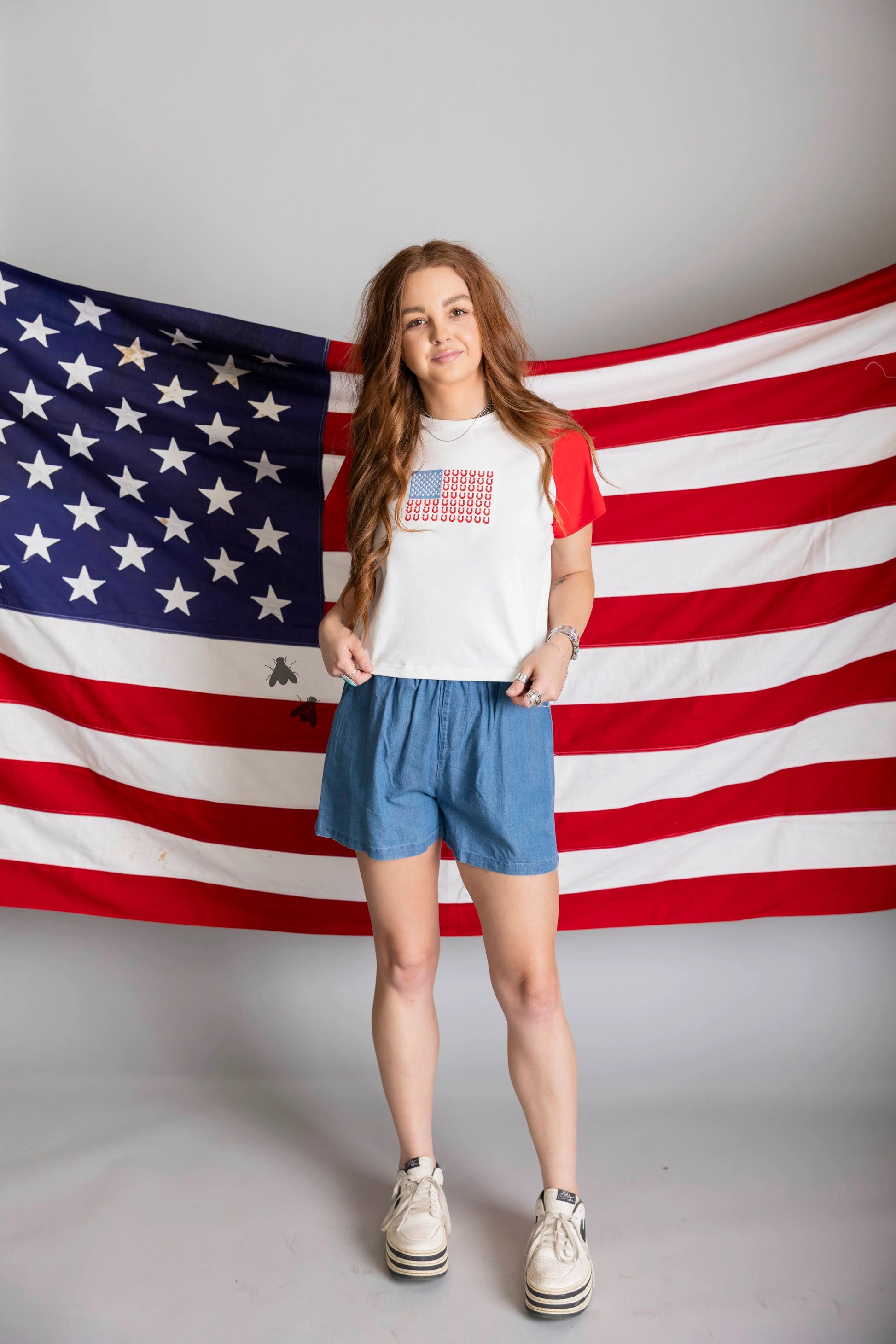 Person wearing a white t-shirt with a graphic design and blue shorts standing in front of an American flag.