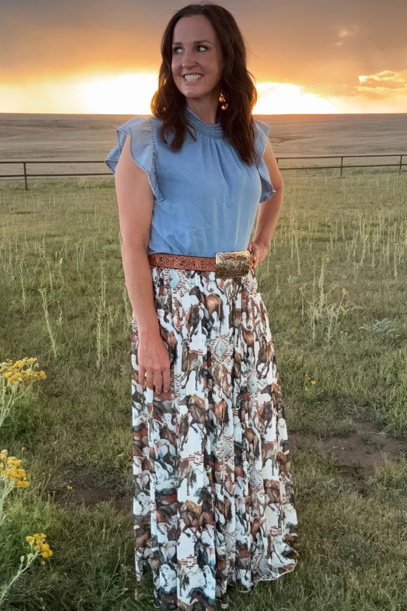 Woman in a blue top and patterned skirt standing in a field with a sunset in the background