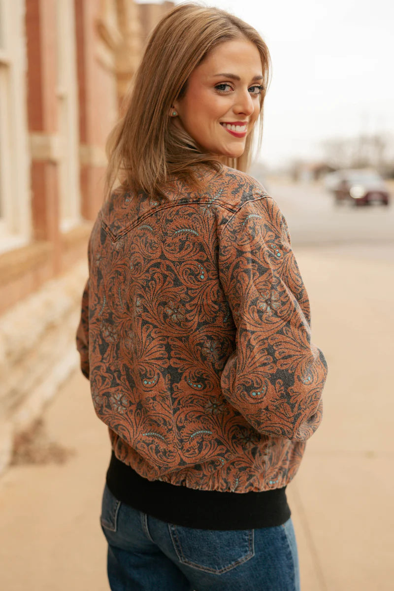 A woman wearing a brown and black paisley patterned bomber jacket, standing outdoors on a street with a blurred background.