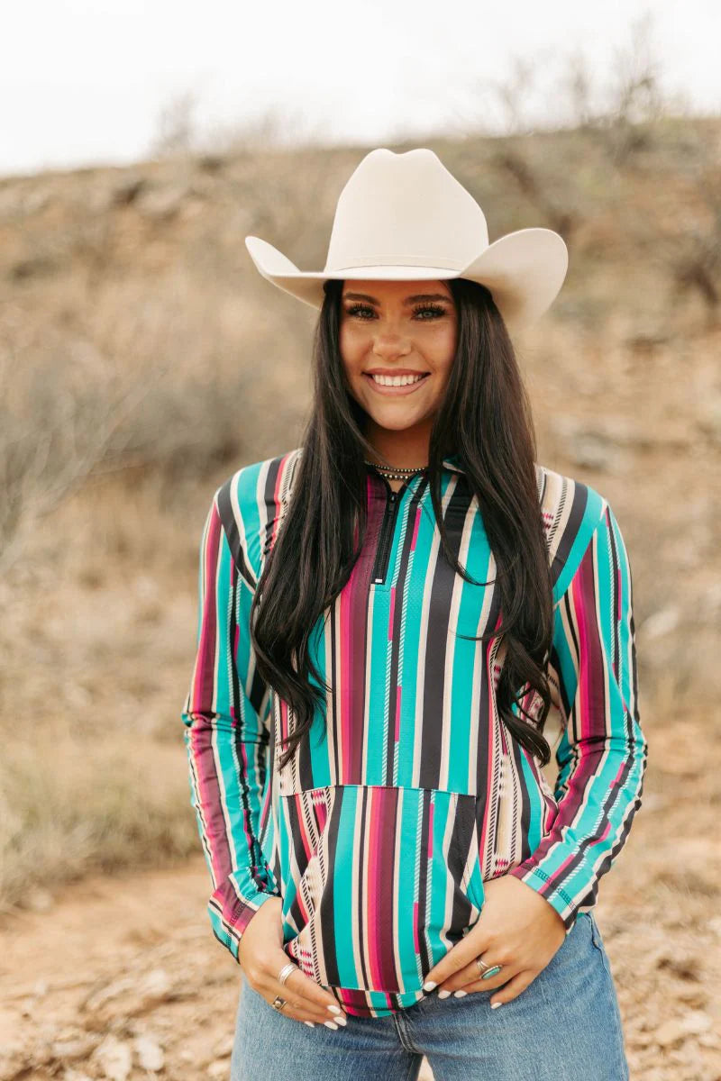 A woman wearing a colorful long-sleeve striped shirt with vertical teal, pink, black, and white stripes, paired with a cream cowboy hat and blue jeans, standing outdoors in a dry, grassy landscape.