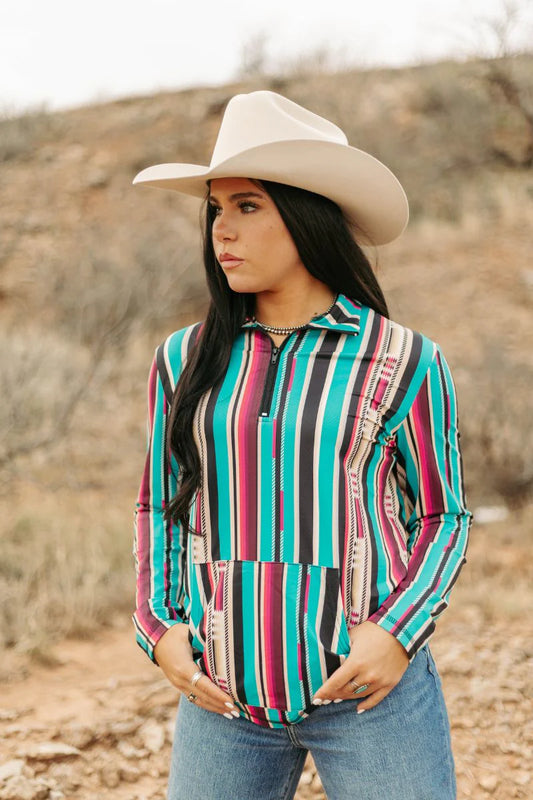 A woman wearing a colorful striped long-sleeve quarter-zip blouse, blue jeans, and a beige cowboy hat, standing in a dry, rocky outdoor landscape.