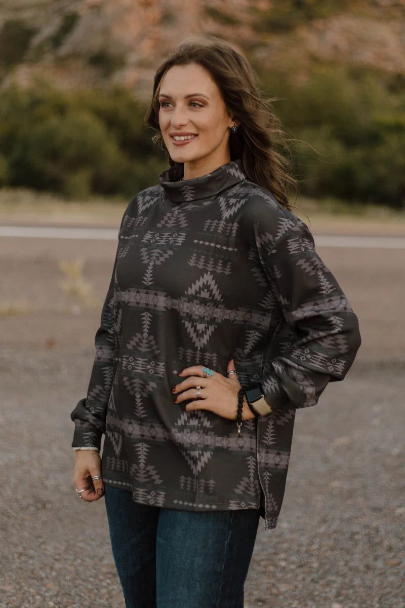 A woman wearing a dark gray and black patterned long-sleeve turtleneck top with blue jeans, standing outdoors on a gravel path with a blurred natural background.