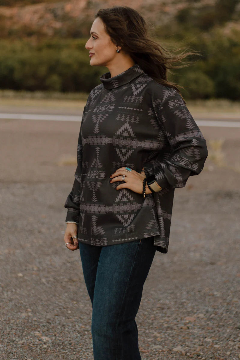 A woman wearing a dark gray patterned long-sleeve mock neck top and dark blue jeans, standing outdoors on a gravel path with a blurred natural background.