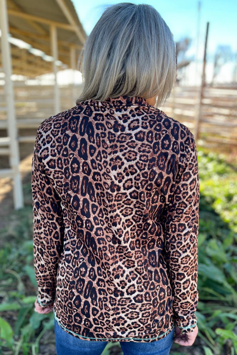 A woman viewed from behind wearing a leopard print long-sleeve top and blue jeans in an outdoor setting with a wooden structure and greenery in the background.