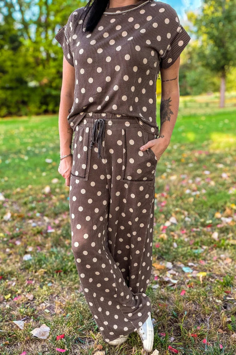 A woman wearing a brown and beige polka dot jumpsuit standing outdoors on a grassy lawn with trees in the background.
