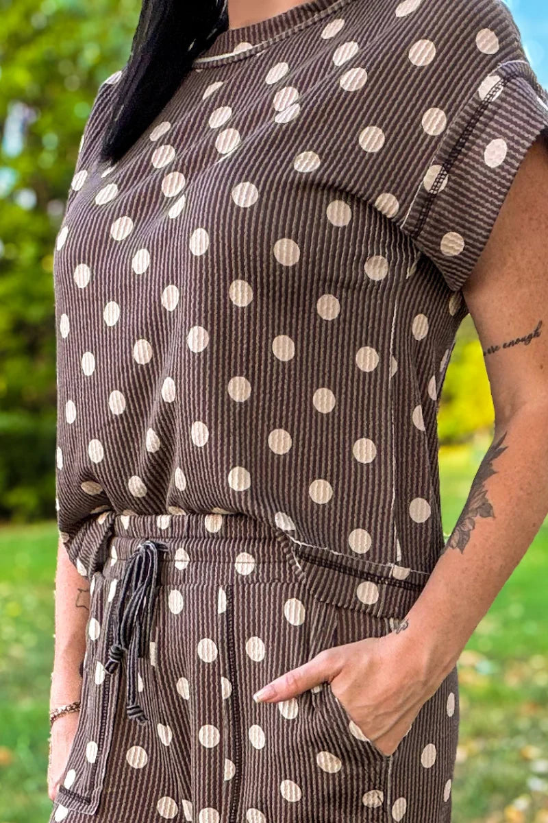 A close-up of a person wearing a brown corduroy jumpsuit with white polka dots, standing outdoors on a grassy lawn.
