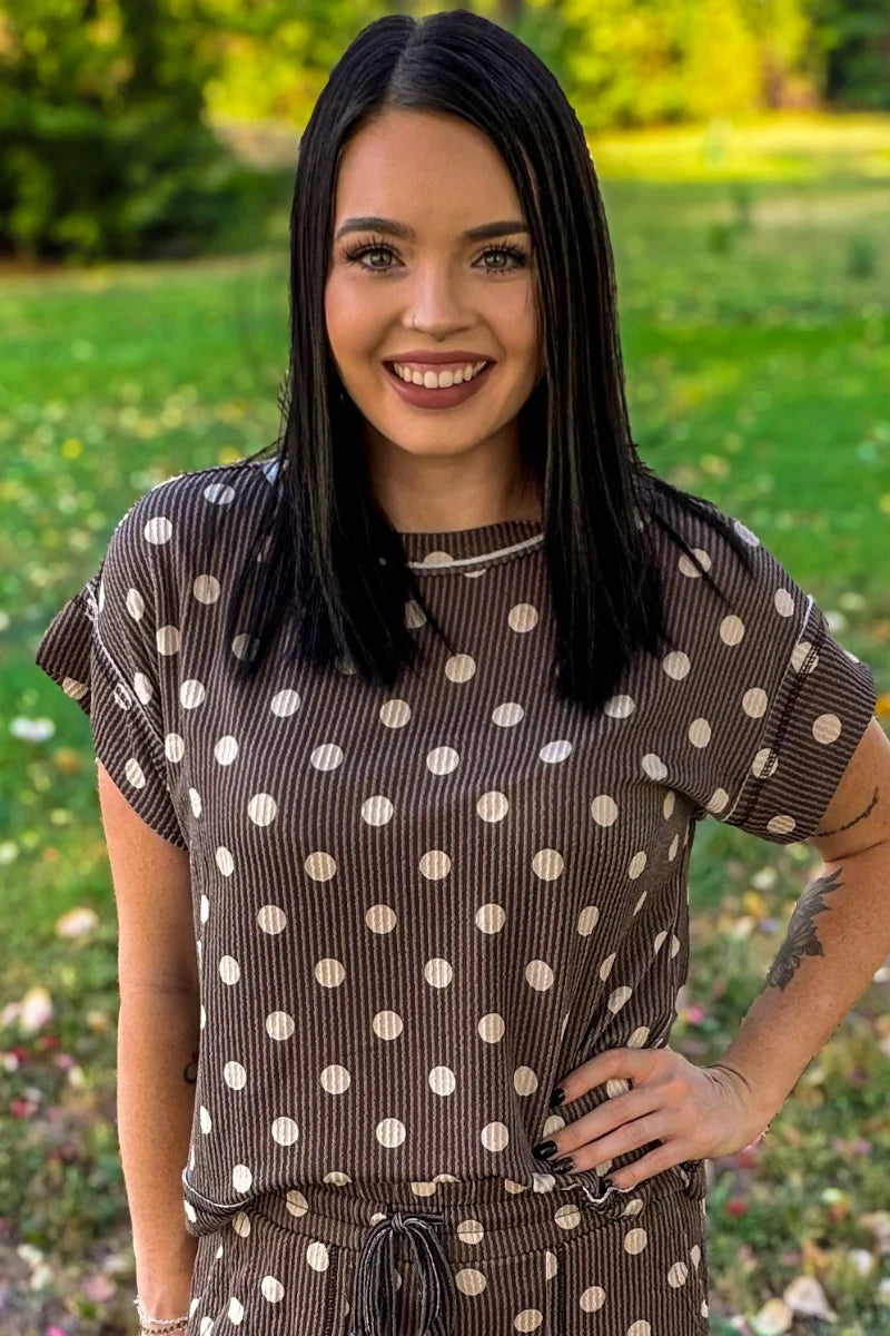 A woman with long black hair smiling outdoors in a brown and white polka dot ribbed top and matching bottoms, set against a blurred green grassy background.