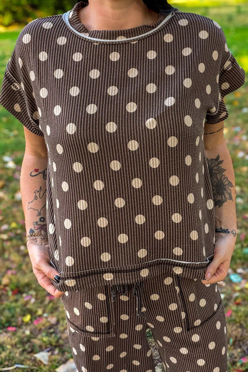 A person wearing a brown ribbed short-sleeve top and matching pants with large beige polka dots, standing outdoors on grass.