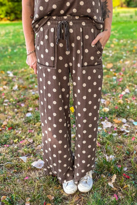 A person wearing brown ribbed wide-leg pants with a cream polka dot pattern, standing outdoors on a grassy lawn with fallen leaves.