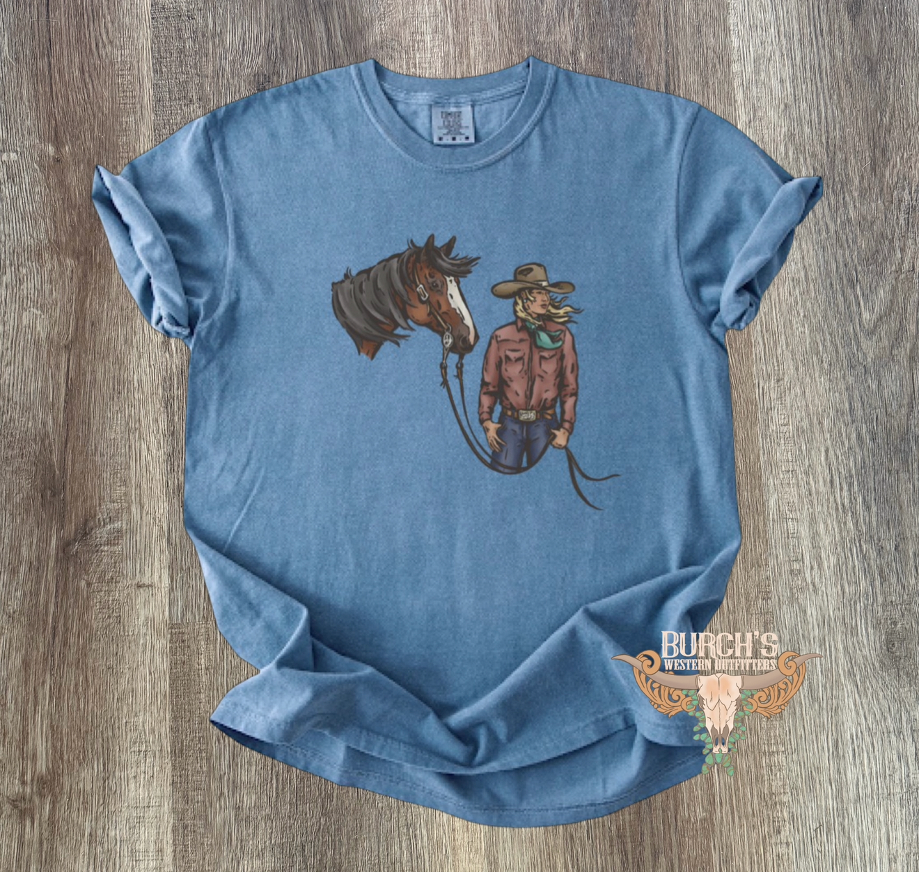 A light blue graphic t-shirt laid flat on a gray wood-textured background. The shirt features a graphic of a brown horse and a cowgirl, and a small logo is printed on the lower right front.