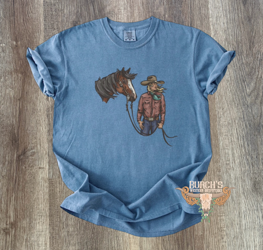 A light blue graphic t-shirt laid flat on a gray wood-textured background. The shirt features a graphic of a brown horse and a cowgirl, and a small logo is printed on the lower right front.
