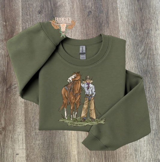 An olive green long-sleeve crewneck sweatshirt with a graphic of a cowboy and his horse, displayed on a rustic wood background.