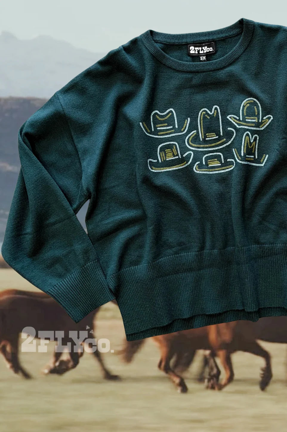 Dark green western sweater featuring embroidered cowboy hat designs in cream and gold on the front, styled against a scenic background with running horses.