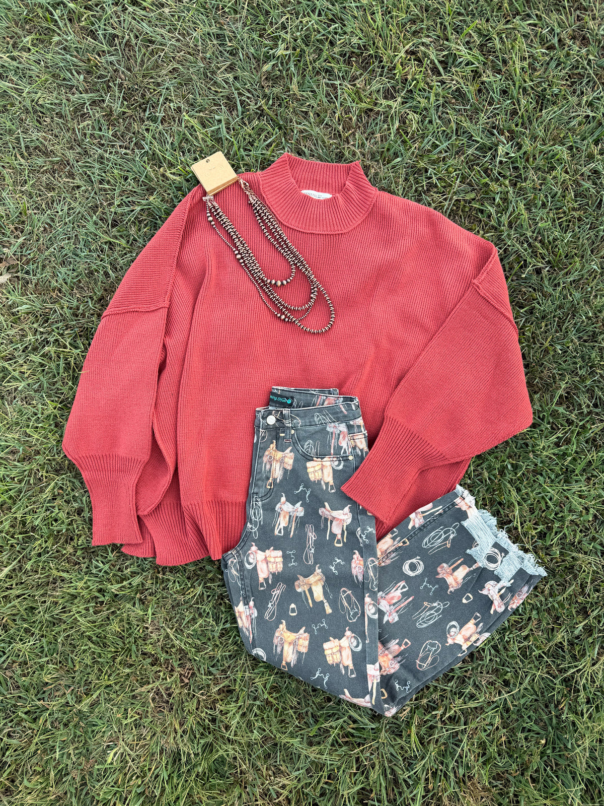 A flat-lay of an outfit on green grass, featuring a coral red knit mock-neck sweater, dark green patterned shorts with a cowboy and cattle motif, and a long dark beaded necklace.