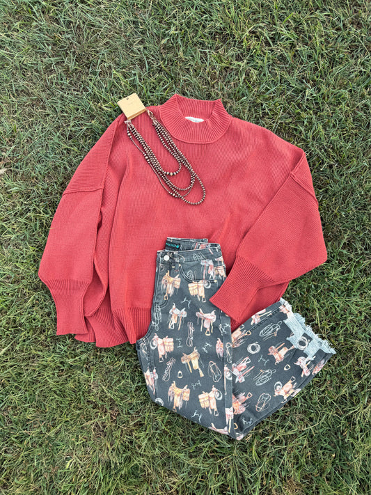 A flat-lay of an outfit on green grass, featuring a coral red knit mock-neck sweater, dark green patterned shorts with a cowboy and cattle motif, and a long dark beaded necklace.